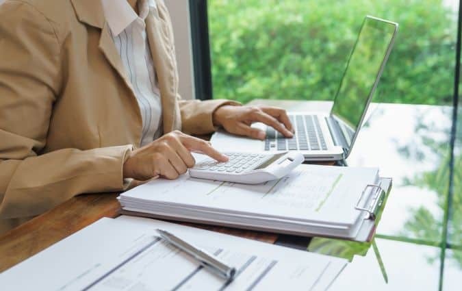 Nő papírok felett számológépen számol, a megtakarításos életbiztosítás költségeit tervezve